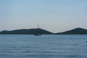 Serene sailboat floating on calm blue sea with lush green hills in the background under clear sky on a sunny day. Concept of tranquility, nature, and nautical adventure.