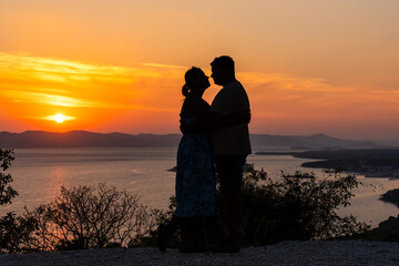 Fototapeta premium Romantic silhouette of couple sharing an embrace during stunning sunset over the sea, with mountains and vibrant orange sky in the background. Concept of love, intimacy, and serene moments in nature.