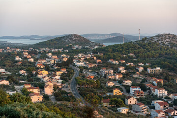 Elevated view of a charming coastal settlement surrounded by lush greenery and winding roads during the golden hour. Concept of tranquil Mediterranean lifestyle, scenic countryside.