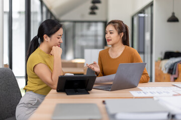 Obraz premium Happy two asian female freelance entrepreneurs reading and planning analyzing the financial report, business plan investment, on computer while working together in the office. 