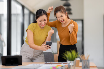 Two asian young women use laptop to work at workplace and lift her arm showing happiness teamwork celebrating achievement and success concept.