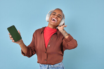 Young African American individual is smiling broadly while wearing headphones. They are immersed in music, holding a smartphone in one hand against a vibrant blue backdrop.