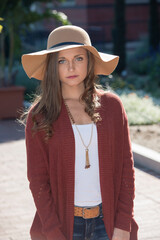 Beautiful young brunette woman in blue jeans, red sweater and wide brimmed hat posing in courtyard