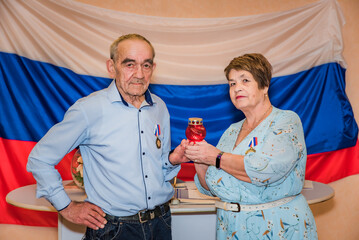 Elderly Russian couple celebrating their golden wedding anniversary in a registry office, standing against a Russian flag backdrop, holding an Orthodox candle symbolizing love and unity