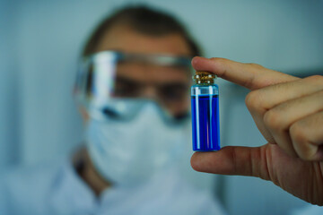 A scientist wearing protective goggles and a face mask holding a vial of blue liquid, closely...