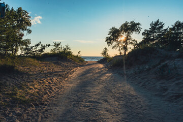 A sandy path bordered by vegetation, leading toward the beach under a clear sky. Sunlight filters...