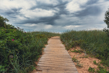 A wooden boardwalk winds through grassy dunes, leading toward the beach under an overcast sky,...