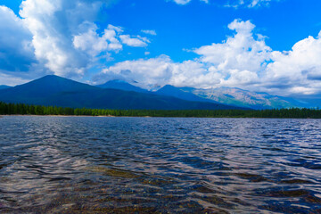 A clear lake with a gently sloping rocky shore, surrounded by lush green forests and distant...
