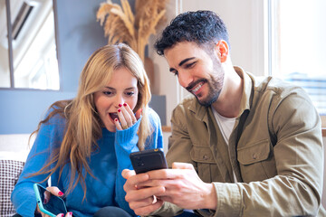 A young couple seated indoors sharing a surprising and joyful moment while looking at a smartphone. The woman covers her mouth in amazement as the man smiles warmly.