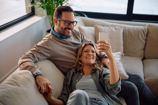 Happy couple using smart phone while relaxing on sofa at home.