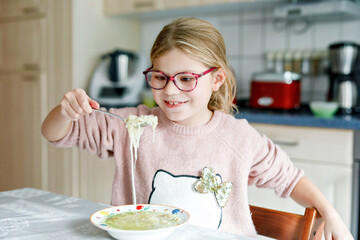 Funny little school girl eat chicken noodles soup with thin pasta. Happy preschool child eating fresh cooked healthy meal with noodles and vegetables at home, indoors.