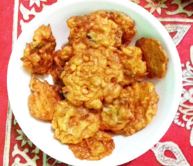 A bowl filled with golden, crispy fritters is placed on a red patterned tablecloth. The fritters appear to be deep-fried, offering a delicious and savory snack option.