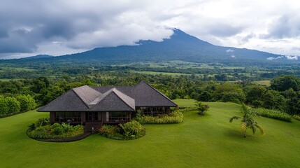 Fototapeta premium A serene aerial view of a modern house surrounded by lush greenery and a majestic mountain backdrop under a cloudy sky.