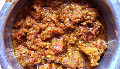 A metal pot is filled with a rich, aromatic curry featuring chunks of tender meat coated in a thick, spicy sauce. The dish appears to be well-cooked with visible oils & vibrant hues of orange & brown.