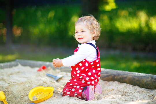 Cute toddler girl playing in sand on outdoor playground. Beautiful baby in red gum trousers having fun on sunny warm summer day. Child with colorful sand toys. Healthy active baby outdoors plays games