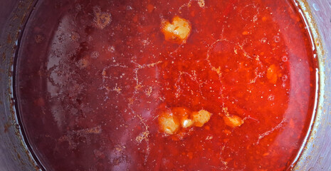 A rich, red liquid with visible oil pools and chunks floats within a pot. The surface has a slight sheen, indicating a hearty sauce or soup.