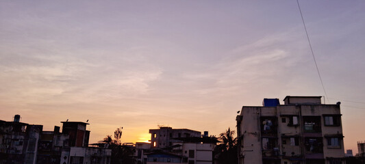 Urban buildings line the foreground silhouetted against a pastel-hued sunset sky. The sun sets near the horizon, casting a warm glow over the cityscape.