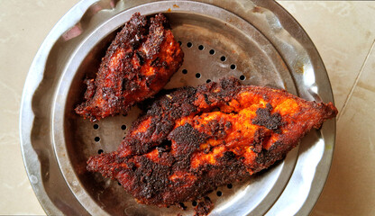 Two pieces of crispy, well-cooked fish are placed on a round metal platter. The fish is richly seasoned, with a dark, spicy crust covering the surface.