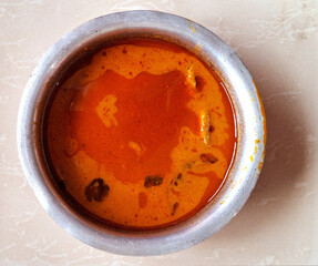 A metal bowl filled with vibrant orange soup sits on a light-colored surface. The soup appears to have a smooth surface with some herbs and spices visible.