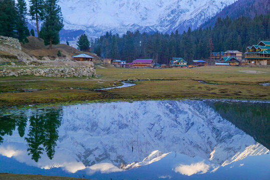Snowy Nanga Parbat reflects perfectly in a calm lake at Fairy Meadows, Pakistan. Surrounded by pine forests, wooden cabins, and a serene meadow.