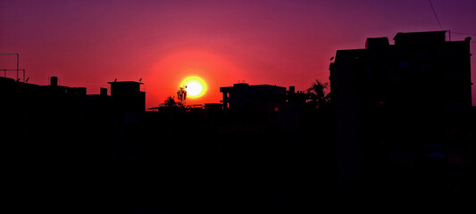 Fototapeta premium A vibrant sunset casts a warm glow over the silhouetted buildings of a cityscape. The sky transitions from deep purple to a bright orange around the sun, creating a dramatic contrast.