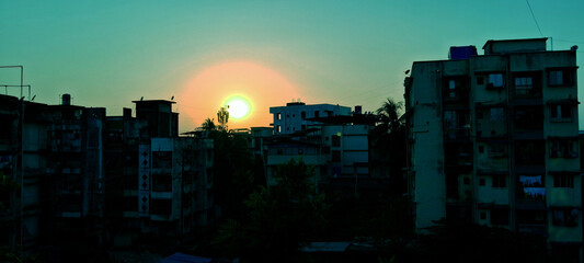 Urban buildings are silhouetted against a glowing sunset, casting a warm green hue across the sky....