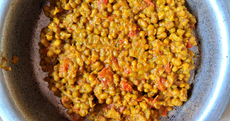 A dish of cooked lentils appears in a metal bowl, seasoned with spices and mixed with chunks of tomato. The vibrant orange and yellow hues suggest a flavorful and aromatic preparation.