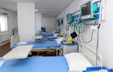 A hospital room with a medical staff member pushing a hospital bed. The room is bright and sterile, with a large window letting in natural light.