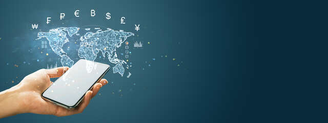 Smartphone in hand displaying global currency symbols and digital map hologram on blue background....