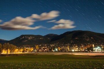Fototapeta premium Serene Night Landscape with Star Trails Over Mountains and Towns Captured in Long Exposure Photograph, Showcasing Tranquil Atmosphere and Natural Beauty