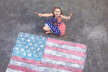 Little girl with glasses and stars and stripes dress, sits on a paved surface beside a large chalk...