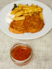 Breaded chicken fillet meal and fried potatoes chips with ridges on a white plate and light color table cloth. Simple food made of high quality premium products. Cooked poultry meat.
