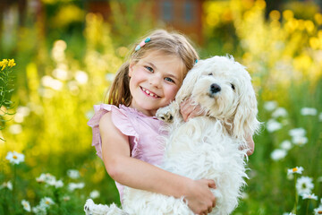 Adorable cute little girl and her puppy on the nature in the summer. Happy school child with eyeglasses holding Maltese dog, having fun with playing. Bright sunset light, active kid.