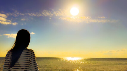 夕暮れ時の夕日とキラキラと光る海を臨む若い女性の後ろ姿