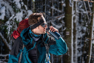 Stylish Man with backpack drinking water from thermos in snowy forest, winter hike