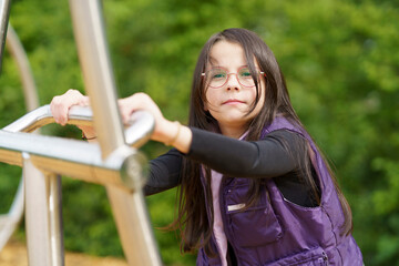 A little cute smiling girl in glasses and a purple vest plays on the playground. Happy childhood...