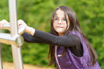 A little cute smiling girl in glasses and a purple vest plays on the playground. Happy childhood...