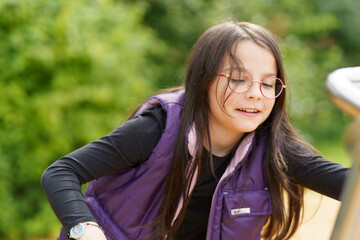 A little cute smiling girl in glasses and a purple vest plays on the playground. Happy childhood...
