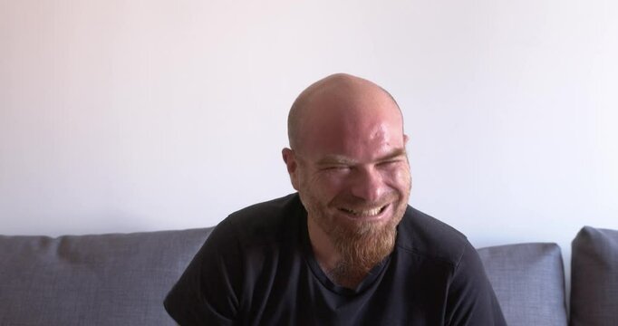Bald man with a red goatee sitting on the couch, laughing heartily and expressing joy, radiating happiness and positive energy.