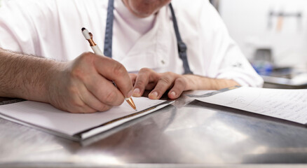 Close-up of a chef in a white uniform writing in a notebook at a stainless steel counter. The scene...