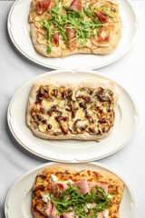 Three delicious flatbreads displayed on white plates. The top features arugula and ham, the middle has cheese and sun-dried tomatoes, and the bottom is topped with ham and fresh greens.