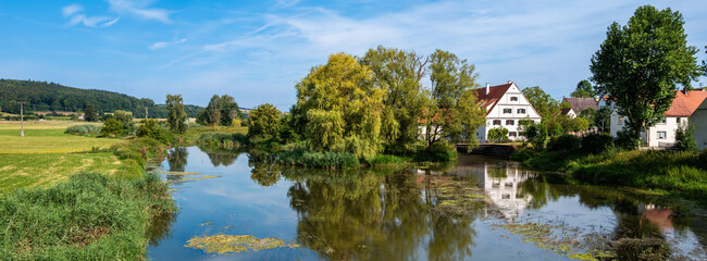 Fototapeta premium A serene landscape featuring a calm Wornitz river reflecting the blue sky and surrounding greenery. Lush trees line the riverbanks, and quaint houses are visible in the background, rural scene