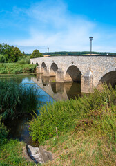 Fototapeta premium Stone bridge with arches over Wornitz river in Germany, surrounded by lush greenery.