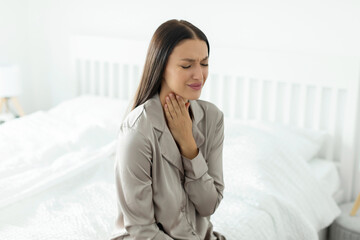 Cough tickling. Sick young woman touching neck sore throat at home, sitting on the edge of bed....
