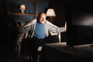 Fan male retired sitting in chair celebrating victory with arms outstretched shouting at screen at night at home.