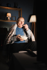 lonely pensioner man sits in a chair at home alone in the evening watching TV, holding a bowl of popcorn in his hands and eating it