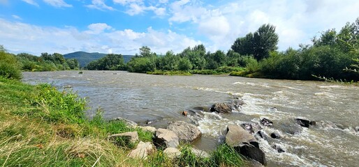 Calm river flows through a lush green landscape under a blue sky with fluffy clouds on a sunny day