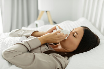 Sick lady suffering from running stuffy nose, lying in bed and blowing her nose using paper napkin tissue. Cold and flu concept