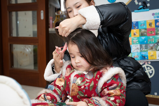 The young Chinese  mother is combing and binding her daughter's hair. Her daughter is wearing traditional Chinese clothes. They're getting ready for celebrating  Chinese New Year.