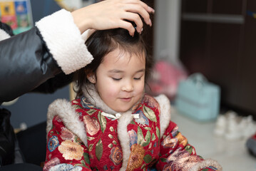 The young Chinese  mother is combing and binding her daughter's hair. Her daughter is wearing...